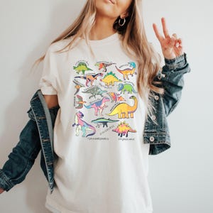 May include: A white t-shirt with a colorful graphic of various dinosaurs. The dinosaurs are outlined in black and filled with different colors. The names of the dinosaurs are written below each dinosaur. The names include: Triceratops, Stegosaurus, Tyrannosaurus, Iguanodon, Hadrosaurus, Pterodactyl, Brachiosaurus, Ankylosaurus, Parasaurolophus, Corythosaurus, and more.