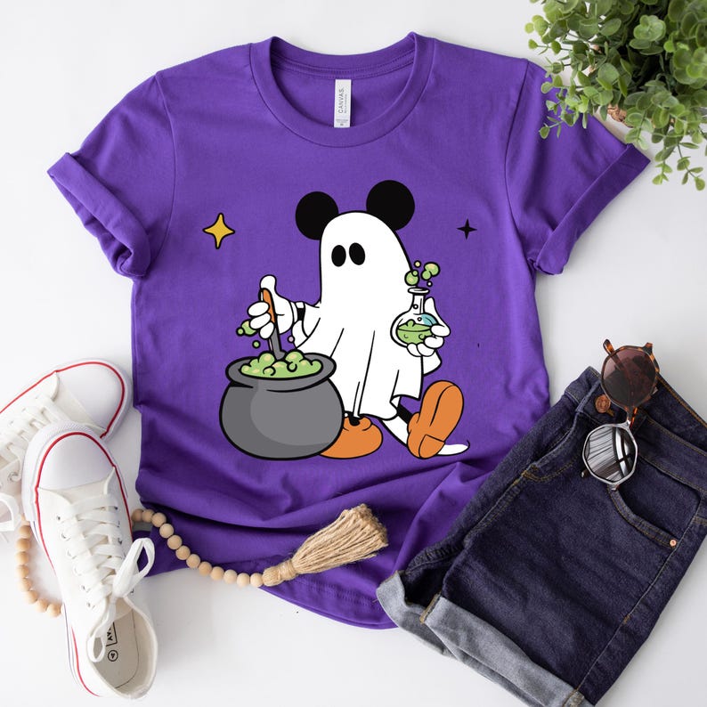 May include: A purple t-shirt featuring a cartoon ghost with Mickey Mouse ears, stirring a cauldron of green potion. The ghost holds a flask with green liquid. The shirt is paired with denim shorts and white sneakers, suggesting a Halloween theme.