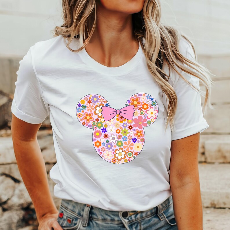 May include: White t-shirt featuring a floral Mickey Mouse design. The design includes a pink bow and the mouse's head and ears are filled with colorful flowers. The shirt is a classic crew neck style.