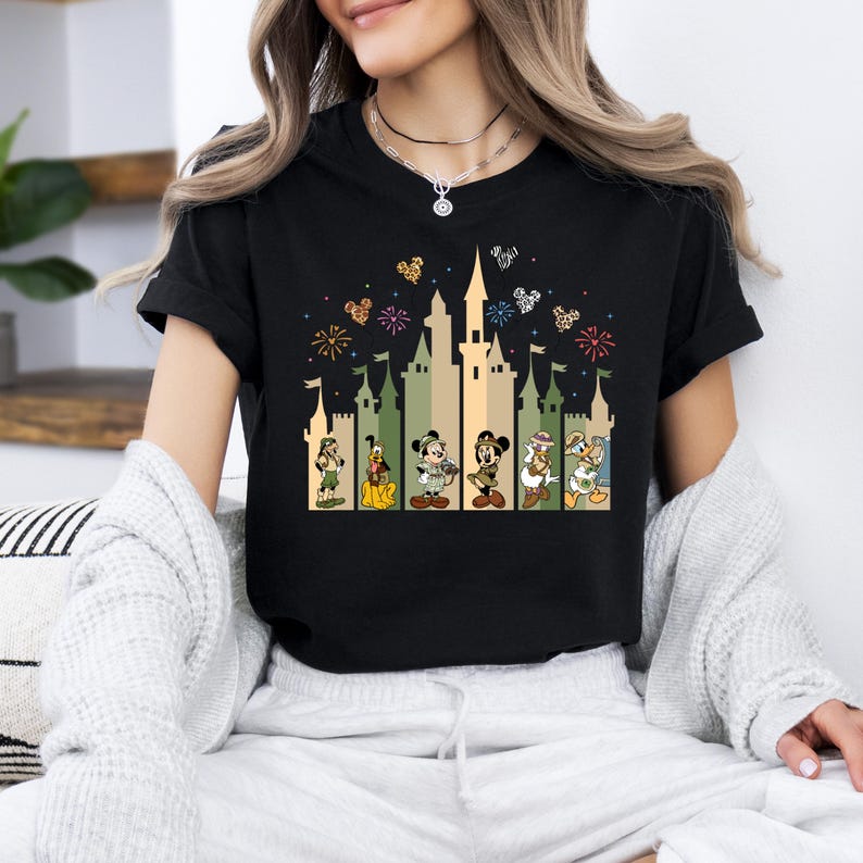 May include: Black t-shirt with a graphic print of the Disney castle and characters. The castle is in a camouflage pattern with fireworks in the background. The characters include Mickey Mouse, Minnie Mouse, Donald Duck, Goofy, Pluto, and Chip and Dale.