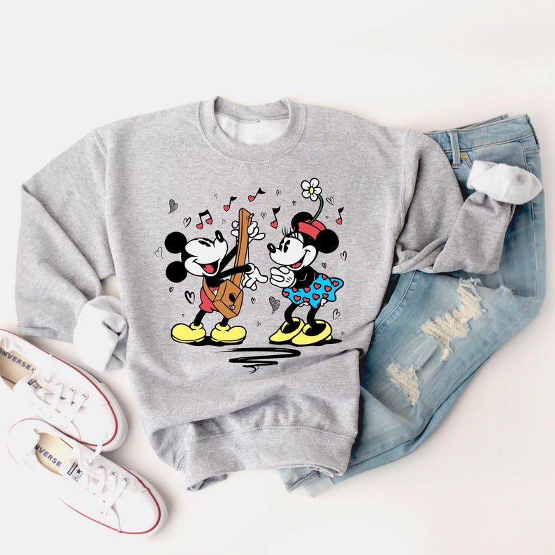 Pode incluir: Uma camisa de moletom cinza com um gr&aacute;fico de Mickey Mouse e Minnie Mouse tocando m&uacute;sica. Mickey est&aacute; tocando guitarra e Minnie est&aacute; segurando uma flor. O gr&aacute;fico &eacute; cercado por cora&ccedil;&otilde;es e notas musicais.