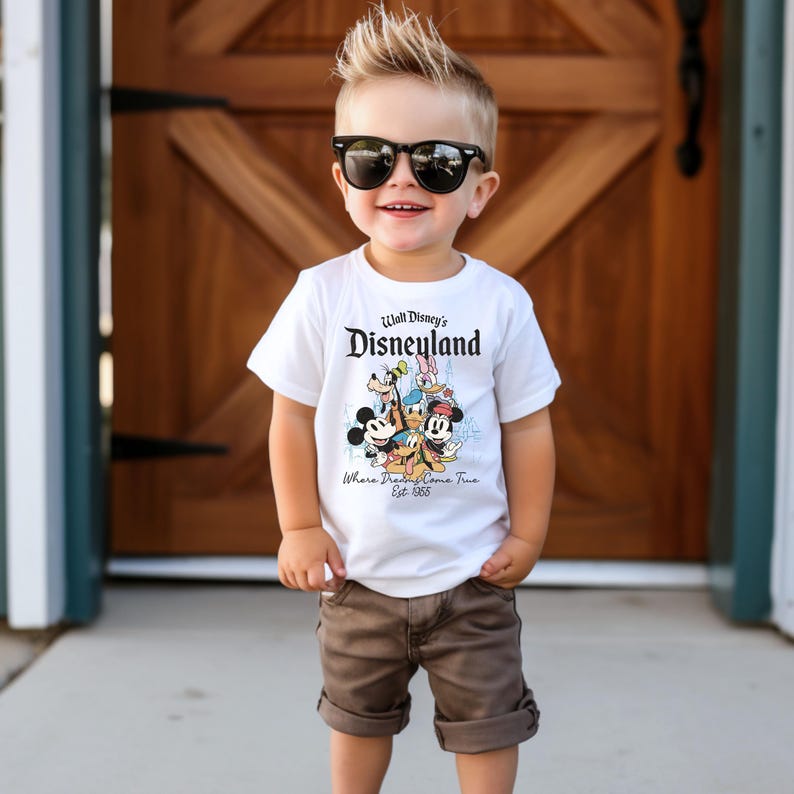 May include: A white t-shirt featuring the Disneyland logo and cartoon characters Mickey Mouse, Donald Duck, Goofy, and others. The shirt is worn by a young child wearing sunglasses and brown shorts.