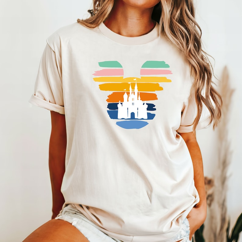 以下が含まれることがあります： ベージュのTシャツに、カラフルなミッキーマウスの頭の中に白いシルエットの城が描かれたグラフィックがあります。グラフィックは、オレンジ、イエロー、ピンク、グリーン、ブルーのストライプで夕焼けを表しています。