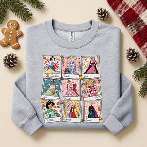 Puede incluir: Una sudadera gris claro con una cuadr&iacute;cula de nueve retratos de princesas de Disney en marcos estilo polaroid. Cada marco tiene un hombre de jengibre y decoraciones navide&ntilde;as. Las princesas son Blancanieves, Cenicienta, Bella, Tiana, Ariel, Aurora, Jasmine, Rapunzel y Elsa.