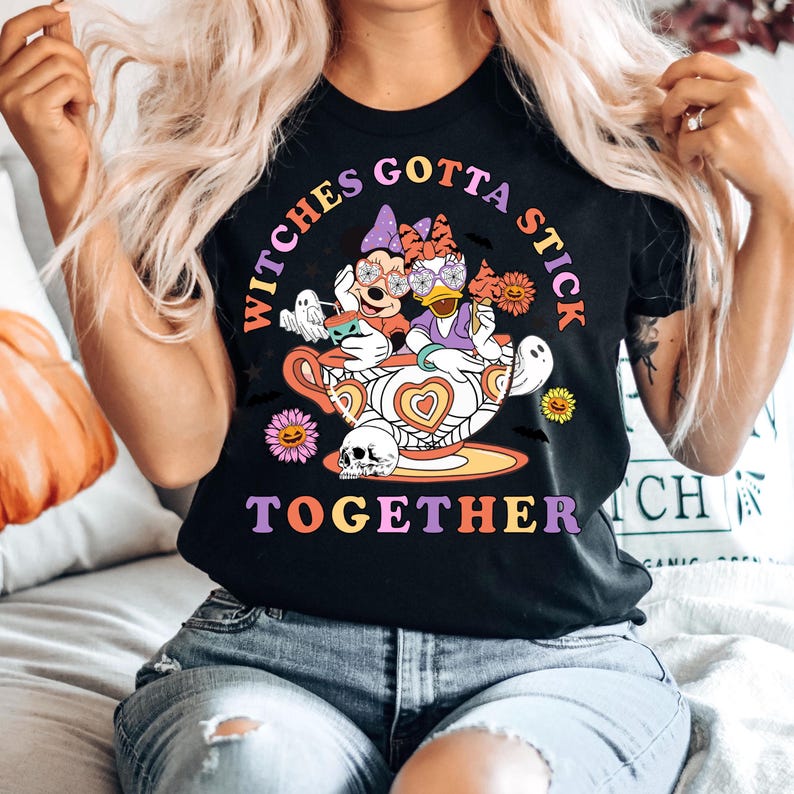 K&ouml;nnte beinhalten: Schwarzes T-Shirt mit einer bunten Grafik von Mickey Mouse und Donald Duck, die als Hexen in einer Teetasse verkleidet sind. Der Text "Witches Gotta Stick Together" ist auf dem Shirt aufgedruckt.