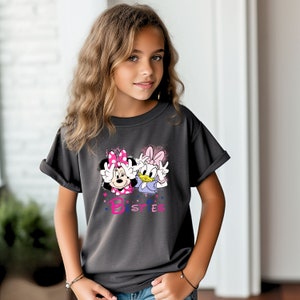 May include: A dark gray t-shirt with a graphic of Minnie Mouse and Daisy Duck. The graphic is in pink, purple, and blue. The text "Bestes" is below the graphic.