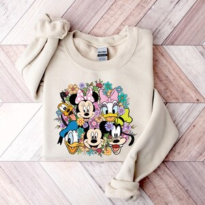 May include: A beige sweatshirt with a graphic print of Mickey Mouse, Minnie Mouse, Donald Duck, Goofy, and Pluto surrounded by flowers.