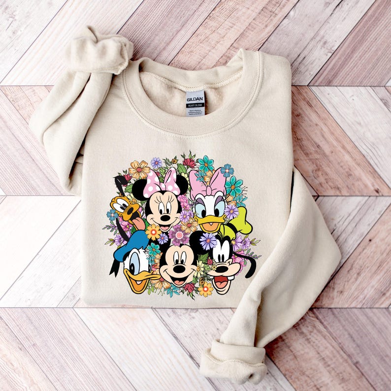 May include: A beige sweatshirt with a graphic print of Mickey Mouse, Minnie Mouse, Donald Duck, Goofy, and Pluto surrounded by flowers.