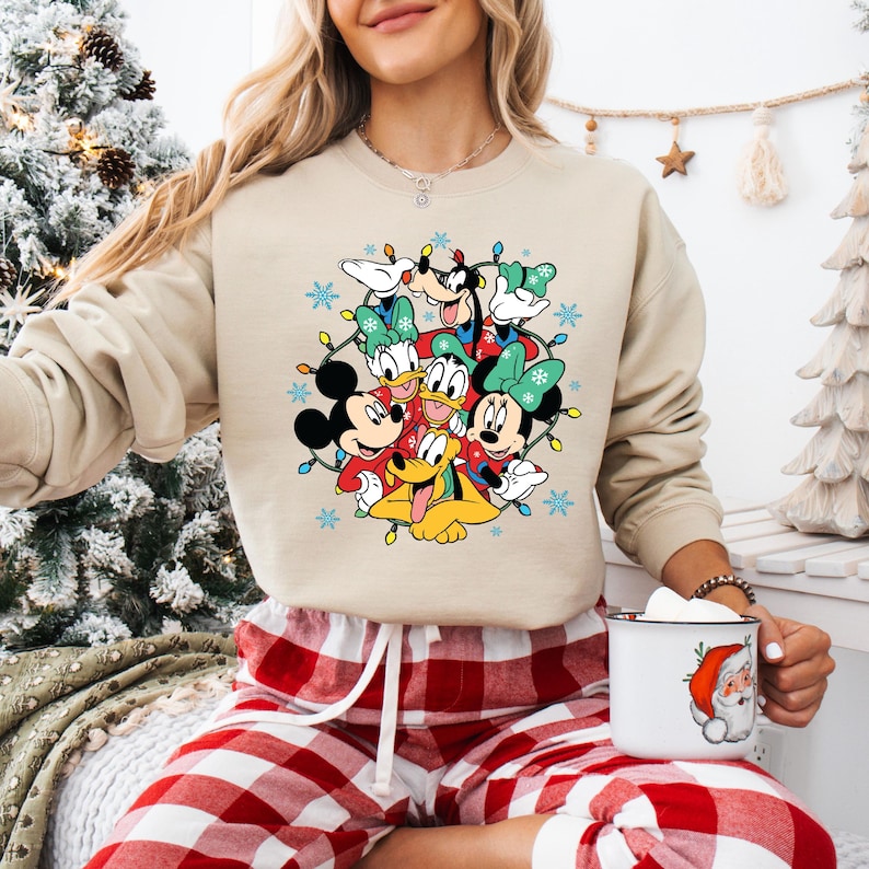 K&ouml;nnte beinhalten: Beiges Sweatshirt mit festlichem Disney-Design. Es zeigt Micky Maus, Minnie Maus, Donald Duck, Goofy und Pluto, umgeben von Weihnachtsbeleuchtung und Schneeflocken. Die Charaktere tragen festliche Kleidung.