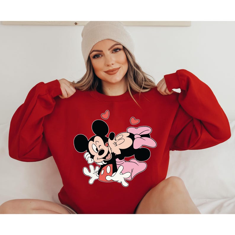 May include: A red sweatshirt featuring a graphic of Mickey and Minnie Mouse kissing, with pink hearts. The cartoon characters are in black, white, and pink. The sweatshirt has long sleeves.