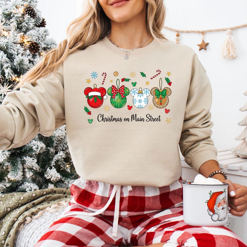 May include: A beige sweatshirt with a Christmas-themed design. It features four Mickey Mouse-shaped ornaments in red, green, and blue, with gingerbread details. The text "Christmas on Main Street" is printed below. The model is wearing red and white plaid pyjama bottoms.