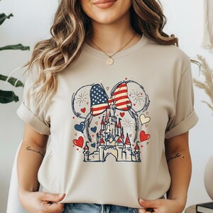 May include: Beige t-shirt featuring a patriotic design. The graphic includes a castle, hearts, and a bow with the American flag pattern. Fireworks and Mickey Mouse ears complete the design.