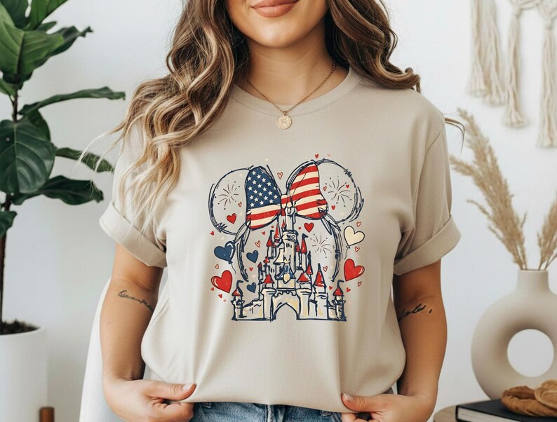 May include: Beige t-shirt featuring a patriotic design. The graphic includes a castle, hearts, and a bow with the American flag pattern. Fireworks and Mickey Mouse ears complete the design.
