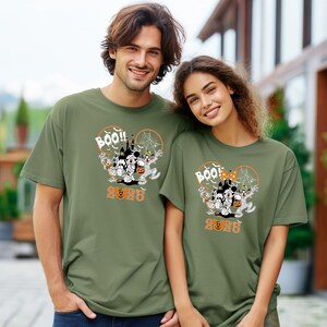 May include: Two people wearing olive green t-shirts with a Halloween graphic. The design features Mickey and Minnie Mouse as mummies, a haunted house, and the word "BOO!!" with the year "2023" in orange.