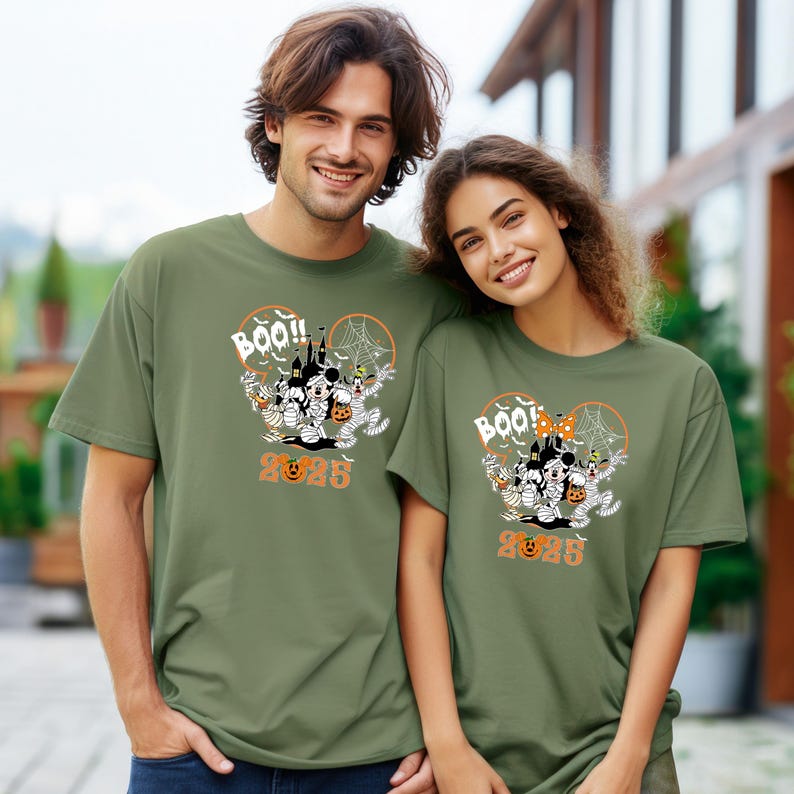 May include: Two people wearing olive green t-shirts with a Halloween graphic. The design features Mickey and Minnie Mouse as mummies, a haunted house, and the word "BOO!!" with the year "2023" in orange.