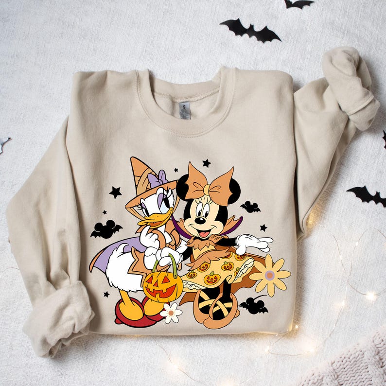 May include: A tan sweatshirt with a Halloween-themed design featuring Minnie Mouse and Daisy Duck dressed in costumes. Minnie is wearing a witch costume with a purple hat and a black dress with orange pumpkins. Daisy is wearing a vampire costume with a red cape and a black dress with orange pumpkins. The design is surrounded by stars and bats.