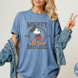 May include: Blue t-shirt featuring a distressed graphic of Mickey Mouse with the text "Mickey Mouse 1928". The shirt is a comfort color blue jean. The shirt is a short-sleeved, crew neck style.