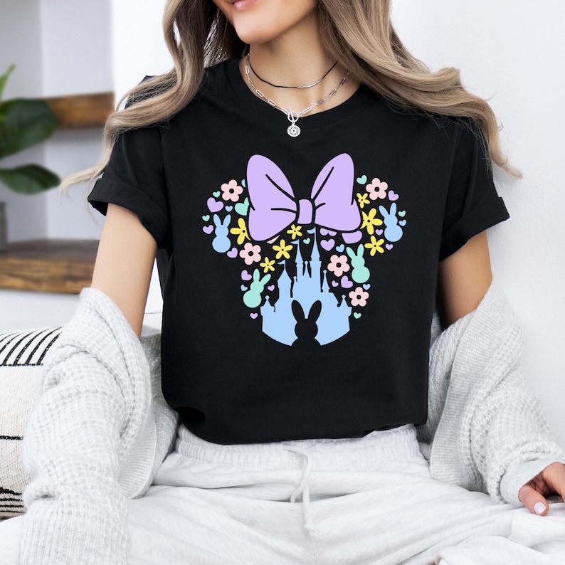 May include: Black t-shirt featuring a pastel-colored design of a bow, flowers, bunnies, and a castle silhouette. The design is reminiscent of a famous cartoon character. The shirt is made of a soft material.