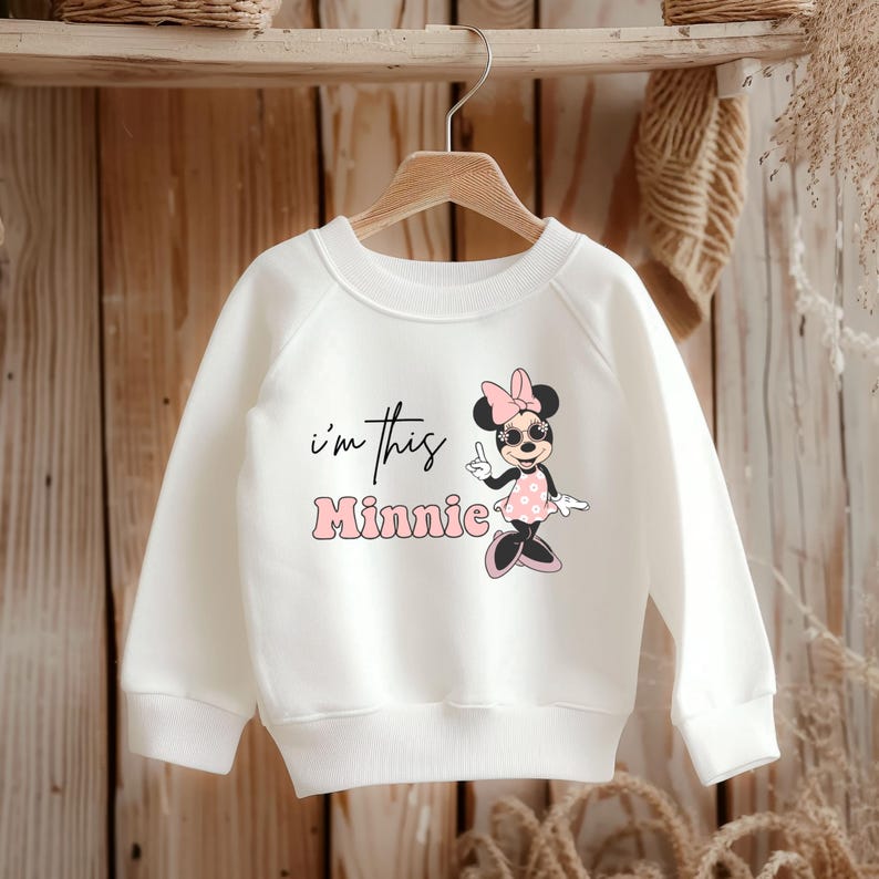 Pu&ograve; includere: Felpa bianca con grafica di Minnie Mouse con occhiali da sole e un vestito rosa a pois. Il testo "I'm this Minnie" &egrave; stampato in nero e rosa. La felpa &egrave; appesa a un gancio di legno.