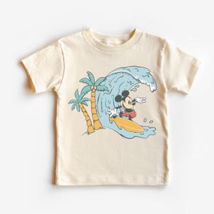 May include: A cream coloured t-shirt featuring a cartoon image of Mickey Mouse surfing on a wave with palm trees in the background.