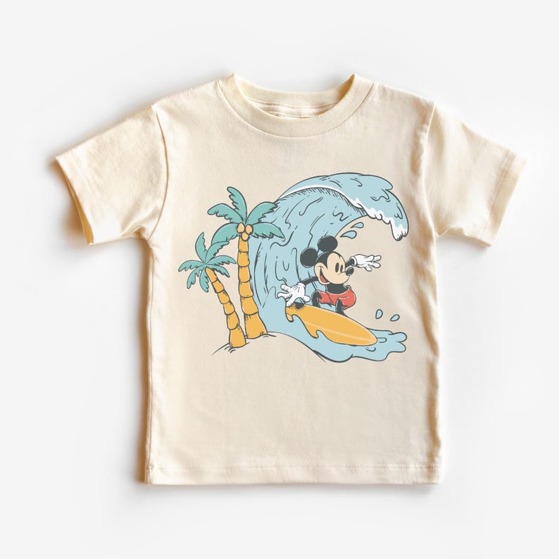 May include: A cream coloured t-shirt featuring a cartoon image of Mickey Mouse surfing on a wave with palm trees in the background.