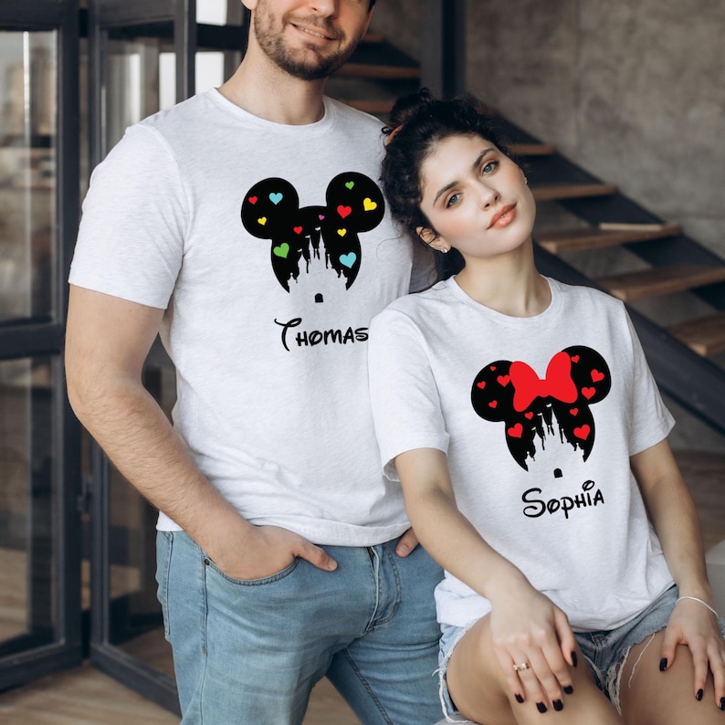 May include: A matching pair of white t-shirts for a couple, featuring black silhouettes of Mickey Mouse and Minnie Mouse heads with a castle inside. The Mickey Mouse shirt has colourful hearts and the text "Thomas" below. The Minnie Mouse shirt has red hearts and a red bow and the text "Sophia" below.