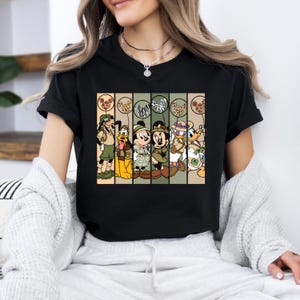 May include: Black t-shirt with a graphic print of Mickey Mouse, Minnie Mouse, Donald Duck, Goofy, and Pluto dressed in safari outfits. The characters are standing in front of a striped background with animal print accents.