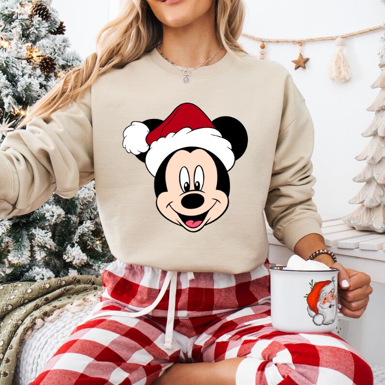 May include: A beige sweatshirt with a Christmas-themed Mickey Mouse graphic. The sweatshirt features a red and white Santa hat on Mickey Mouse's head. The person is wearing red and white plaid pajama pants.