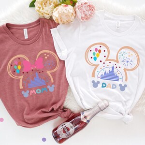 May include: Two t-shirts with a Mickey Mouse ear design featuring a pink bow, a purple castle, and colorful balloons. The text "MOM" and "DAD" are printed on the shirts.