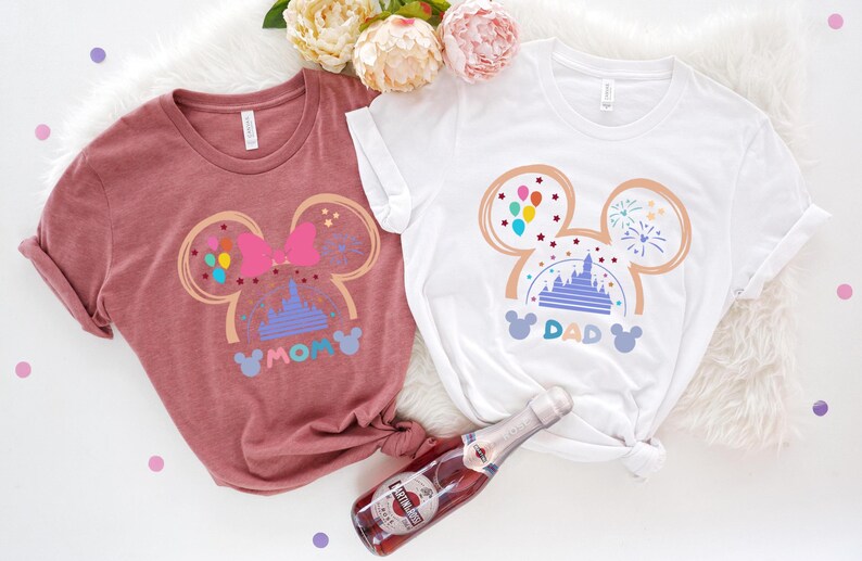 May include: Two t-shirts with a Mickey Mouse ear design featuring a pink bow, a purple castle, and colorful balloons. The text "MOM" and "DAD" are printed on the shirts.