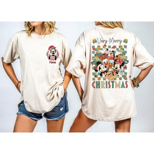 May include: Beige t-shirt featuring a front graphic of Goofy in a Santa hat and the name "Nurse". The back of the shirt displays a "Very Merry Christmas" design with Disney characters, Christmas trees, and gift boxes. The word "CHRISTMAS" is printed at the bottom.
