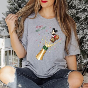 May include: A grey t-shirt with a cartoon of Mickey Mouse celebrating the New Year, popping a champagne bottle. The design includes the text "Happy New Year!" with gold, green, and red accents, creating a festive theme.