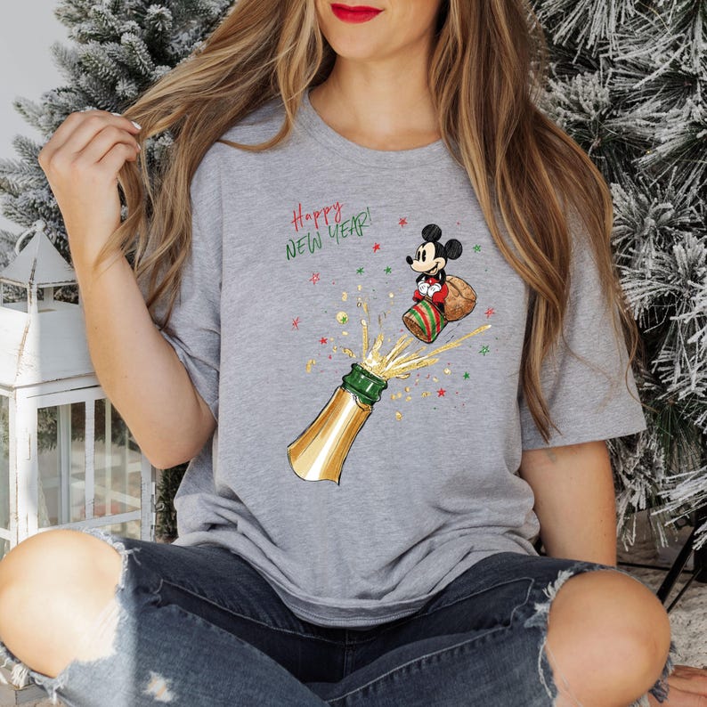 May include: A grey t-shirt with a cartoon of Mickey Mouse celebrating the New Year, popping a champagne bottle. The design includes the text "Happy New Year!" with gold, green, and red accents, creating a festive theme.