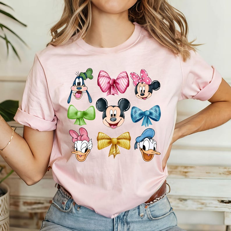 May include: Pink t-shirt with a graphic of Disney characters wearing bows. The characters include Mickey Mouse, Minnie Mouse, Donald Duck, Goofy, and Daisy Duck.