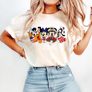 Peut inclure: Un t-shirt blanc avec un imprim&eacute; graphique de Mickey Mouse, Minnie Mouse, Donald Duck, Dingo et Pluto portant des tenues de marin. Les personnages sont tous souriants et ont des accents rouge, blanc et bleu.