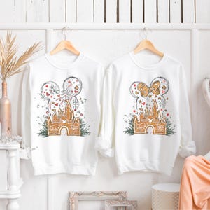 K&ouml;nnte beinhalten: Zwei wei&szlig;e Sweatshirts mit langen &Auml;rmeln, jeweils mit einem Mickey Mouse-Kopf-Design. Eines hat ein Feuerwerks- und Schlossdesign, das andere eine Schleife und ein Schloss. Das Schloss ist im Lebkuchen-Stil gehalten, mit gr&uuml;nen Akzenten.