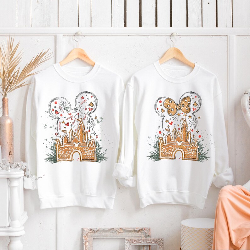 K&ouml;nnte beinhalten: Zwei wei&szlig;e Sweatshirts mit langen &Auml;rmeln, jeweils mit einem Mickey Mouse-Kopf-Design. Eines hat ein Feuerwerks- und Schlossdesign, das andere eine Schleife und ein Schloss. Das Schloss ist im Lebkuchen-Stil gehalten, mit gr&uuml;nen Akzenten.