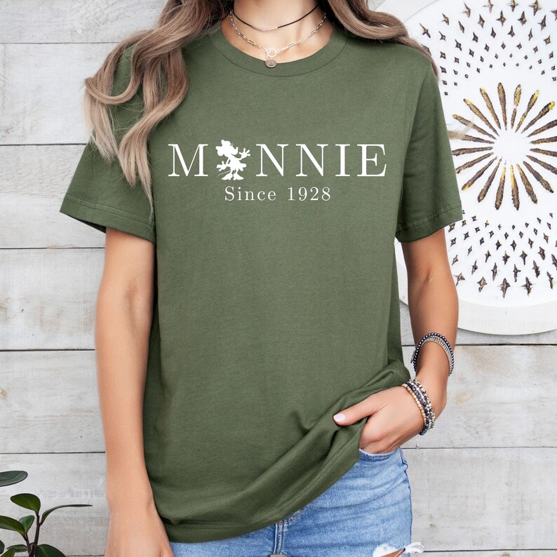 May include: Olive green t-shirt with the word "Minnie" in white, featuring a cartoon character graphic, and the text "Since 1928". The shirt has short sleeves and a crew neck.
