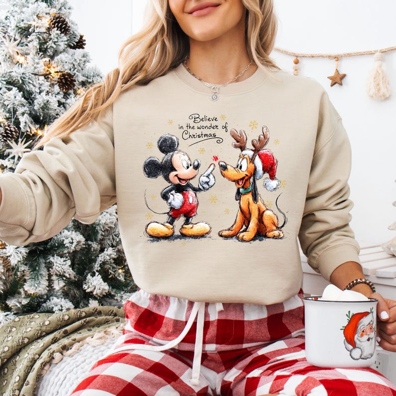 May include: Beige sweatshirt featuring Mickey Mouse and Pluto with the text "Believe in the wonder of Christmas." Mickey Mouse is wearing a red shirt and Pluto is wearing a Santa hat. The sweatshirt is paired with red and white plaid pajama pants.