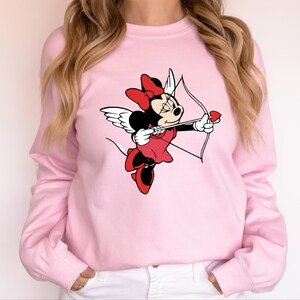May include: Pink sweatshirt featuring a graphic of Minnie Mouse dressed as Cupid, holding a bow and arrow with a heart-shaped arrowhead.