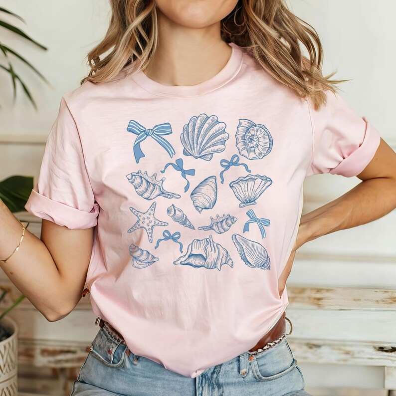 Pu&ograve; includere: Una maglietta rosa con un design di conchiglie blu e bianche. Il design presenta varie conchiglie, stelle marine e fiocchi.