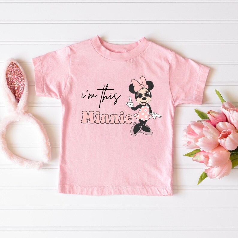 Pu&ograve; includere: Una maglietta rosa chiaro con la scritta "I'm this Minnie" e una grafica di Minnie Mouse. La grafica di Minnie Mouse indossa occhiali da sole e un vestito a pois. Un paio di orecchie da coniglio rosa e un mazzo di fiori rosa sono anche nell'immagine.