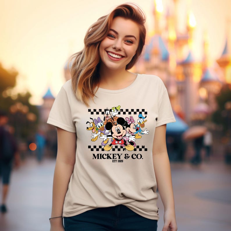 May include: A beige t-shirt featuring a colorful graphic of Mickey Mouse and friends, including Donald Duck, Goofy, and Minnie Mouse. The design is set against a black and white checkered pattern, with the text "MICKEY & CO. EST 1928" below.