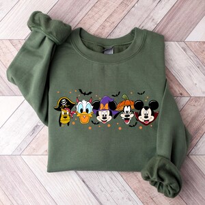 Op de afbeelding: Groen sweatshirt met een Halloween-thema ontwerp met Mickey Mouse, Donald Duck, Goofy en Pluto verkleed. De personages dragen hoeden en accessoires zoals een piratenhoed, een heksenhoed, duivelshoorns en een pompoen.