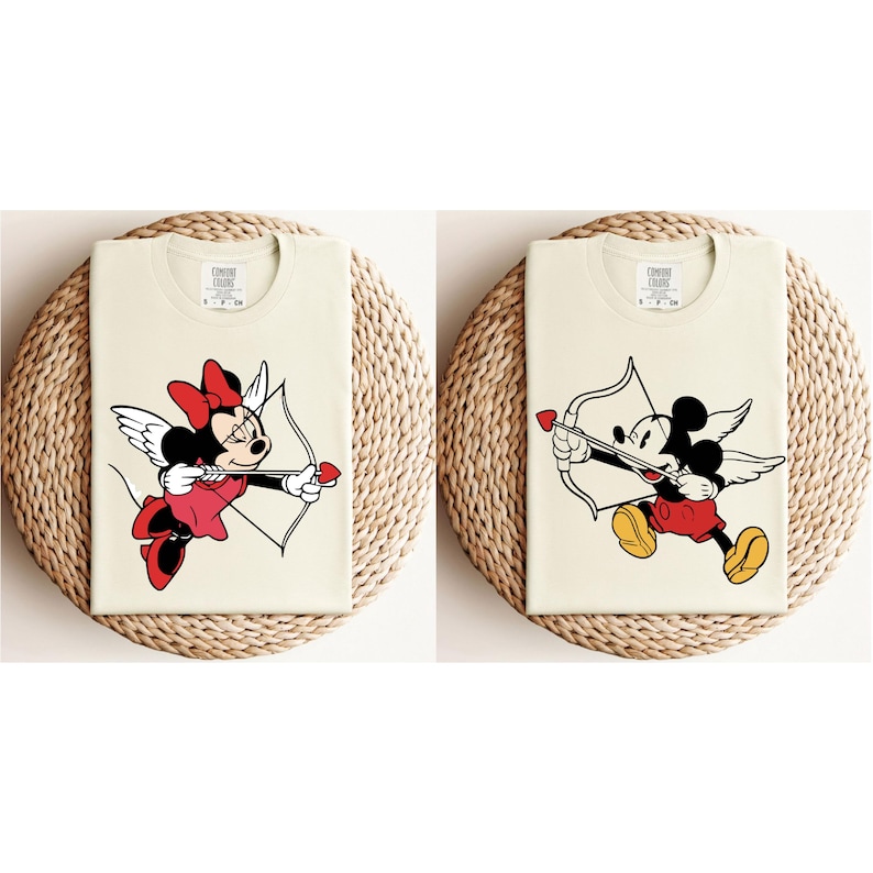 May include: Two cream-coloured t-shirts featuring a cartoon image of Mickey Mouse and Minnie Mouse dressed as cupids. Mickey Mouse is wearing red shorts and wings, holding a bow and arrow. Minnie Mouse is wearing a red dress and wings, holding a bow and arrow.