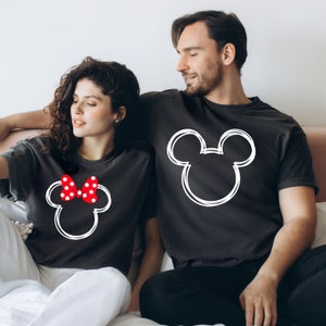 May include: A couple wearing matching black t-shirts with a white outline of Mickey Mouse's head. The woman's shirt has a red bow with white polka dots on Mickey's head.