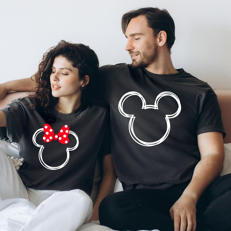 May include: A couple wearing matching black t-shirts with a white outline of Mickey Mouse's head. The woman's shirt has a red bow with white polka dots on Mickey's head.