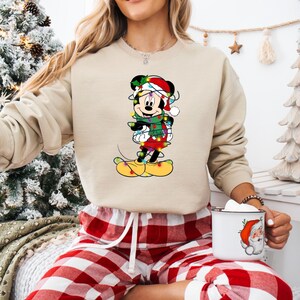 K&ouml;nnte beinhalten: Beiges Sweatshirt mit Rundhalsausschnitt und einem festlichen Mickey Mouse-Motiv. Mickey tr&auml;gt einen Weihnachtsmannhut, einen gr&uuml;nen Schal und eine Lichterkette. Die Person tr&auml;gt eine rot-wei&szlig; karierte Pyjamahose und h&auml;lt eine Tasse mit einem Weihnachtsmann-Design.