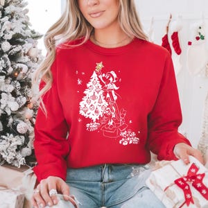 May include: Red Christmas sweatshirt with a white graphic of Mickey Mouse decorating a Christmas tree. The graphic includes a star, ornaments, and presents.