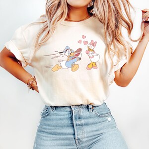 Op de afbeelding: Een wit T-shirt met een cartoon afbeelding van Donald Duck die rent met een boeket bloemen en Daisy Duck die met gekruiste armen staat. De achtergrond is wit met roze harten.
